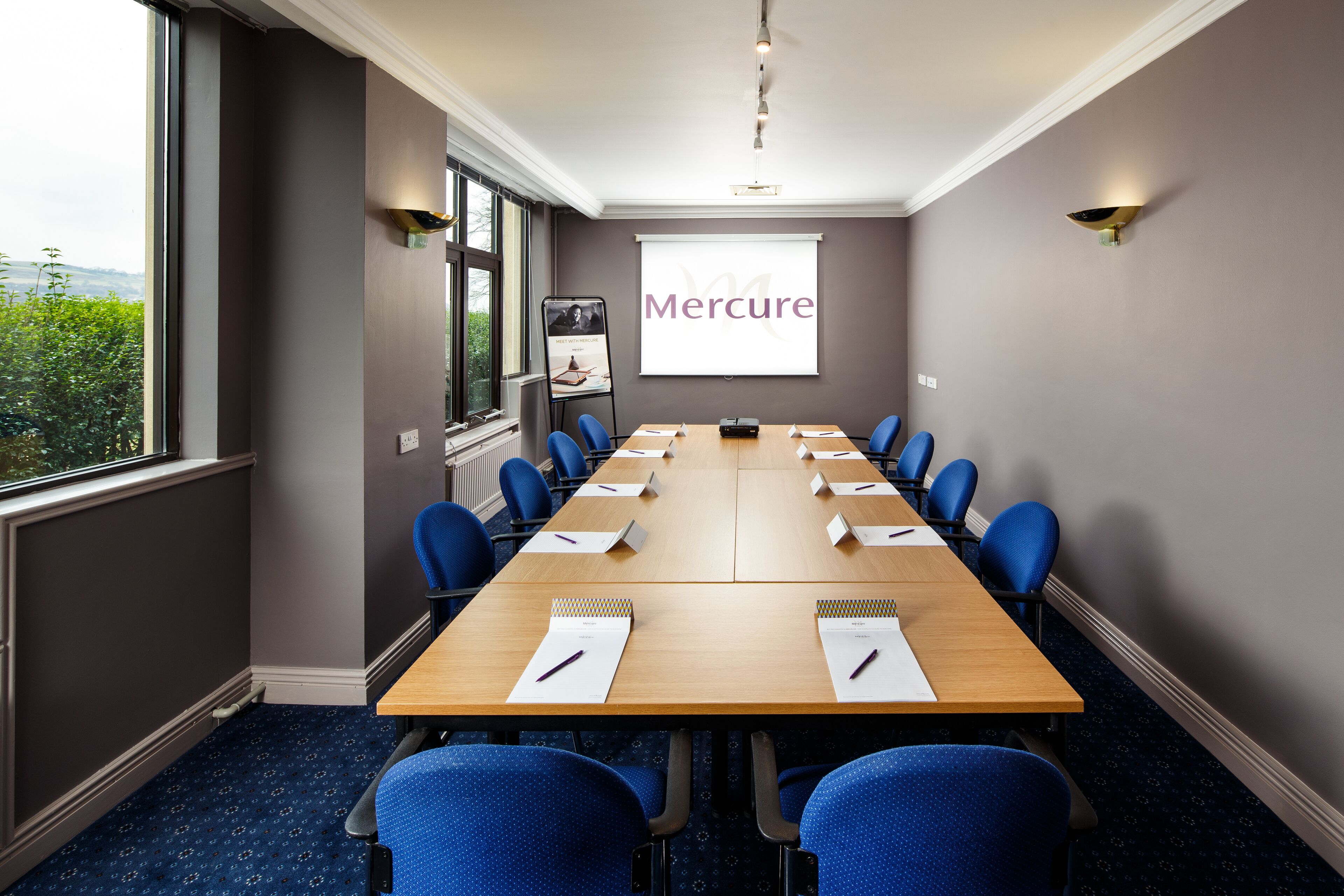 A conference room with a long table, blue chairs, and a projector screen displaying "Mercure".