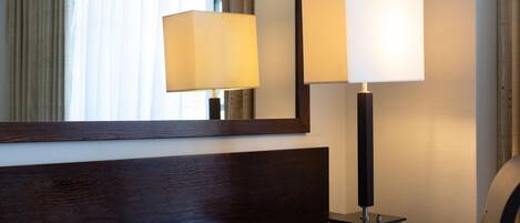 A hotel room with a wooden headboard, a bedside table, a lamp, and a mirror reflecting the room.