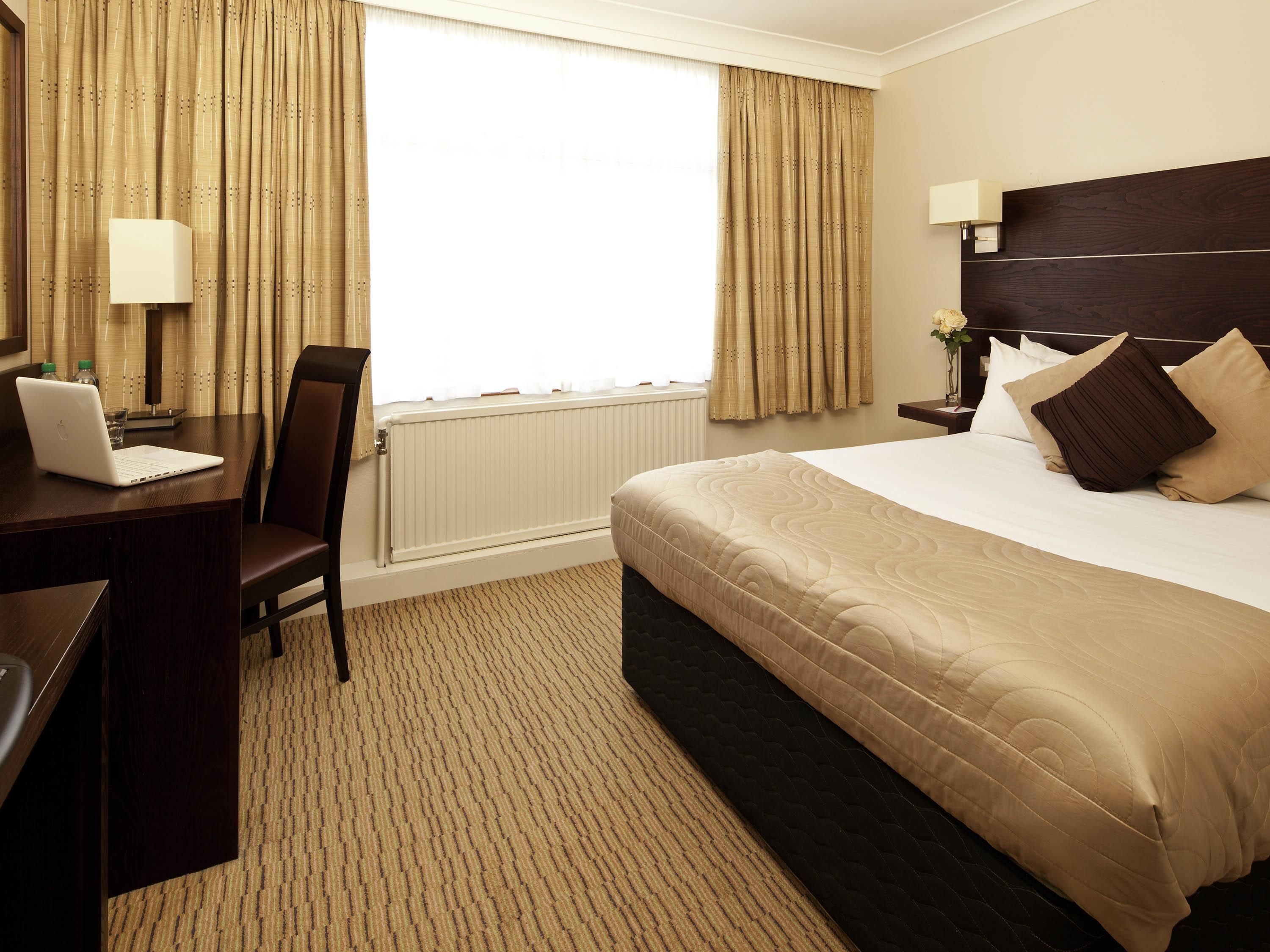 Habitación de hotel con cama, escritorio con computadora portátil, silla, mesita de noche con lámpara y cortinas.