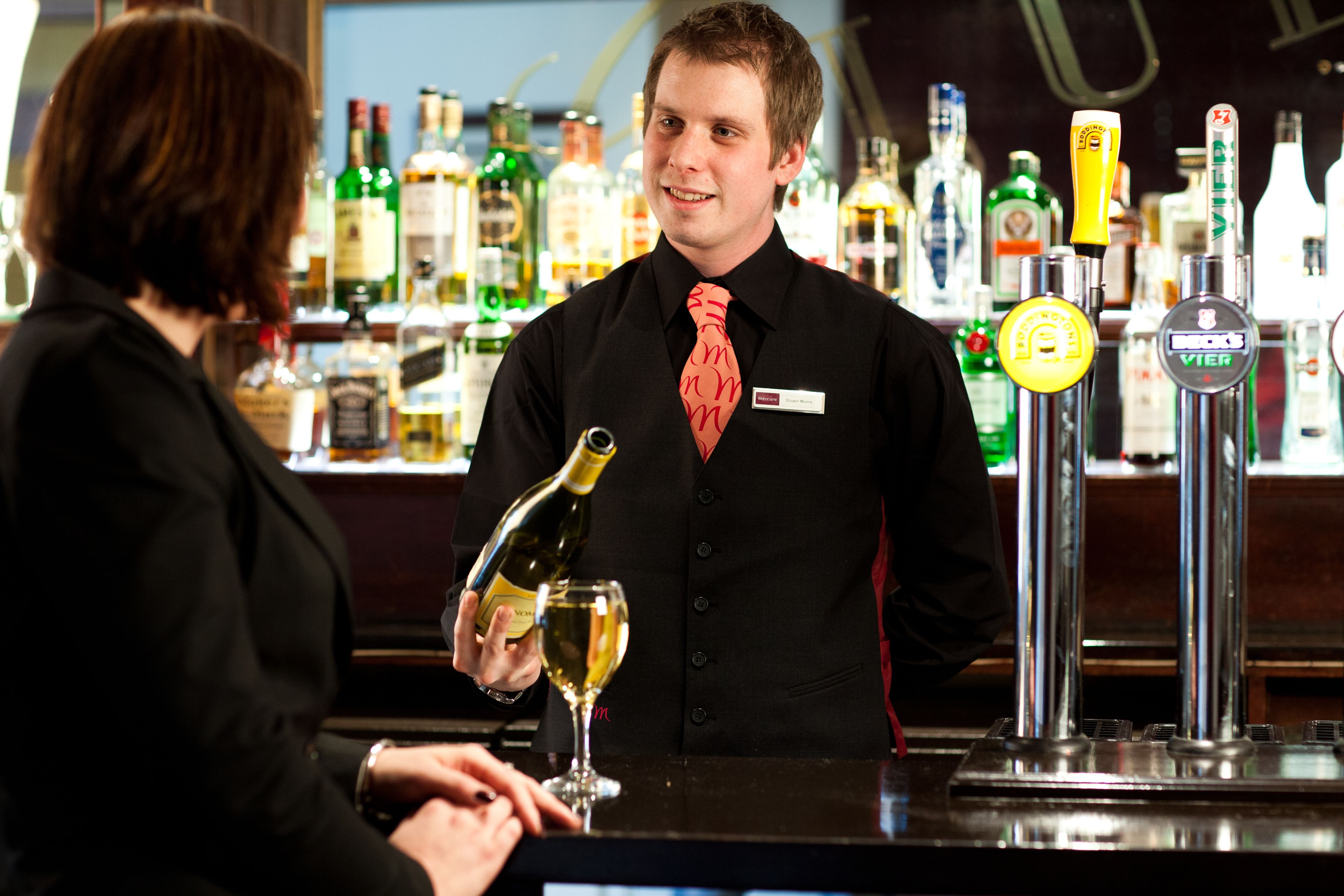 A bartender pouring a drink into a glass at a bar with various bottles in the background.