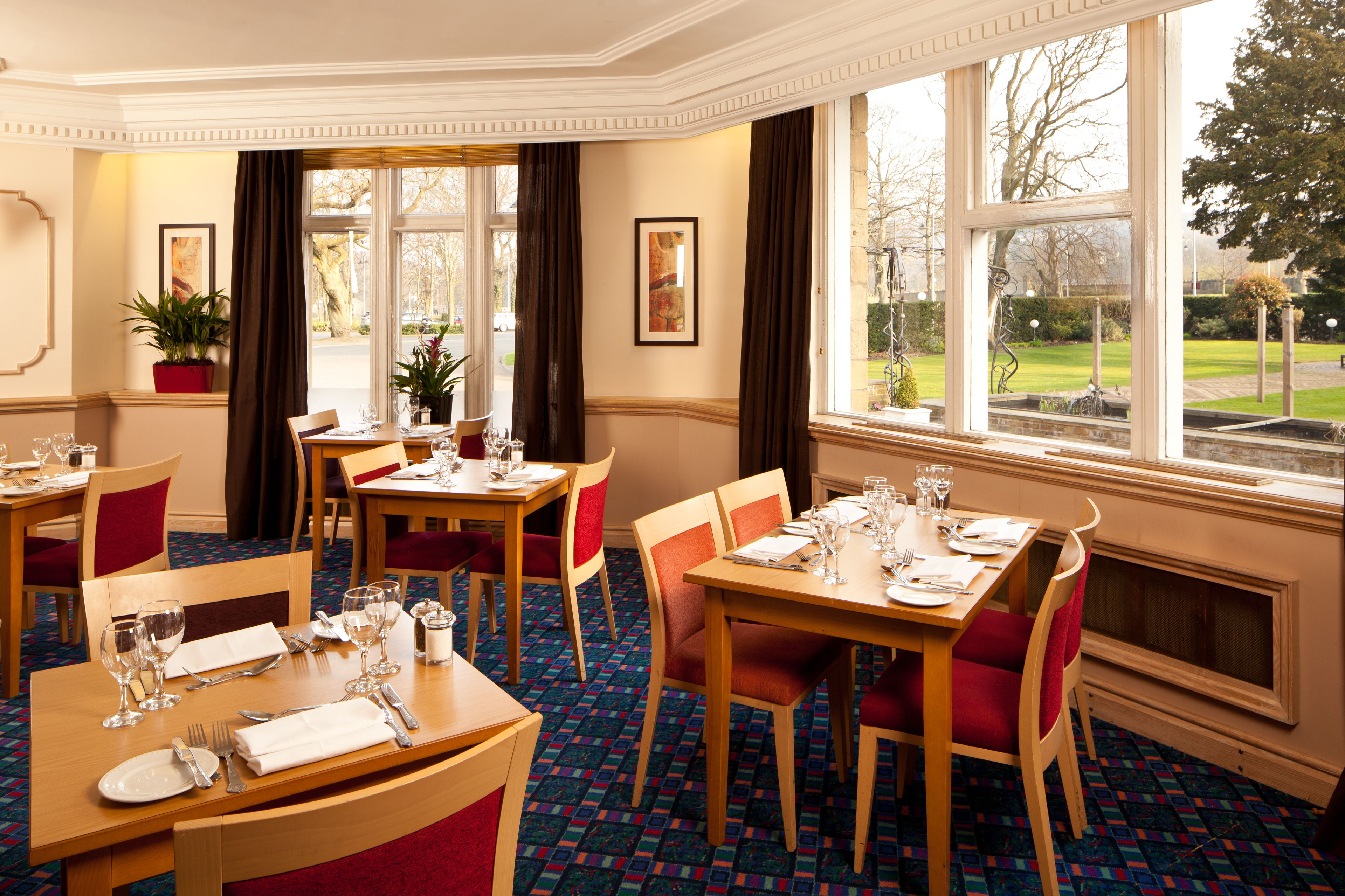 A hotel dining area with multiple tables set for guests, large windows offering a view of the outdoors, and a decorative plant in the corner.