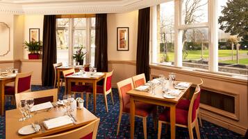 A hotel dining area with multiple tables set for guests, large windows offering a view of the outdoors, and a decorative plant in the corner.