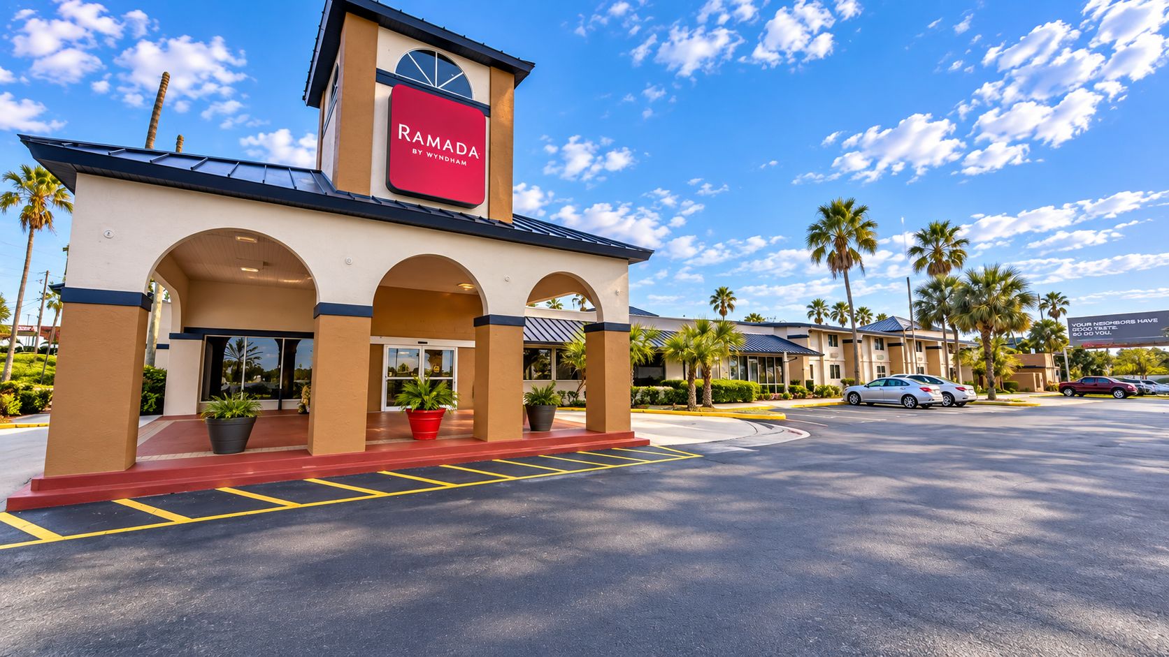 Un hôtel Ramada au design moderne, caractérisé par une enseigne rouge bien visible, une entrée couverte avec des arcades et un parking où sont stationnées plusieurs voitures.