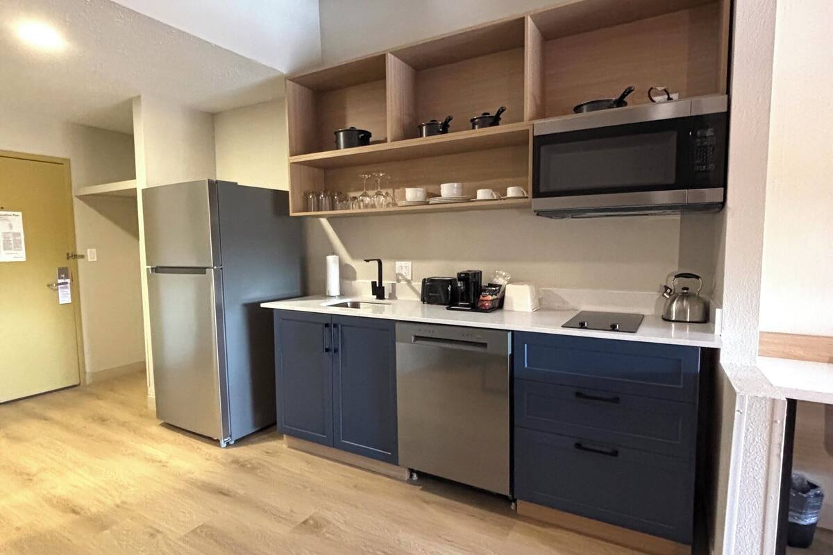 A modern kitchen with a stainless steel refrigerator, microwave, and dishwasher. There are wooden shelves above the counter and a small sink area.