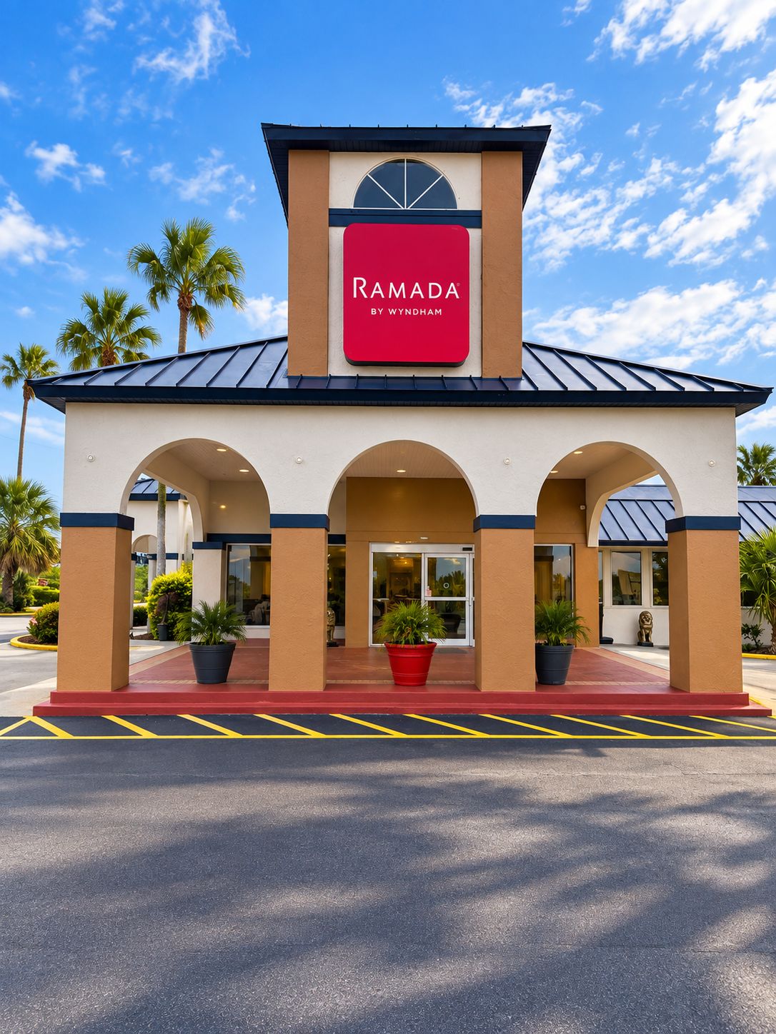 Un hotel Ramada con un letrero rojo, una entrada techada y palmeras al fondo.