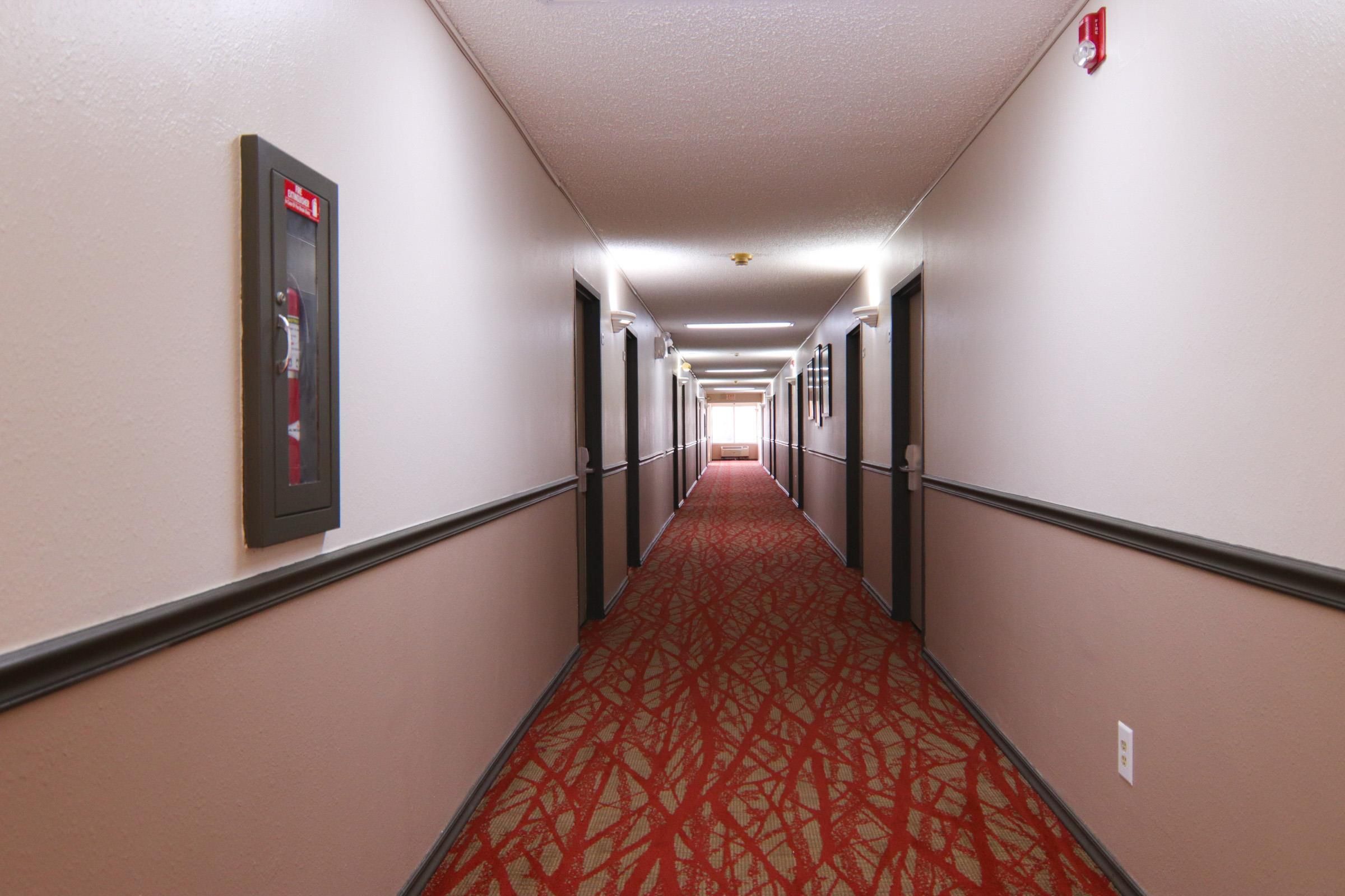 A hotel hallway with a red patterned carpet, doors on both sides, and a fire alarm on the left wall.