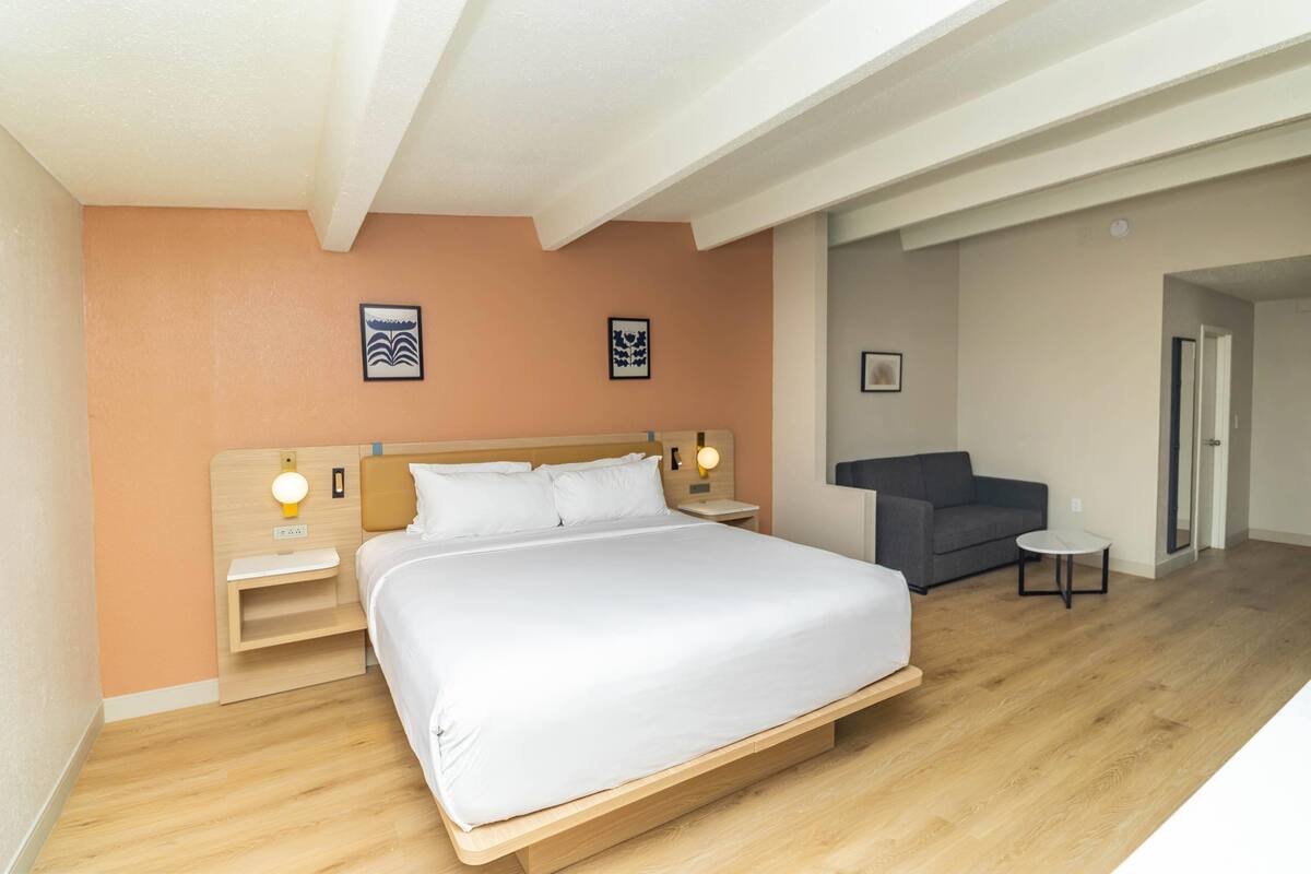 A hotel room with a large bed, a nightstand with a lamp, a small table, a sofa, and two framed pictures on the wall.