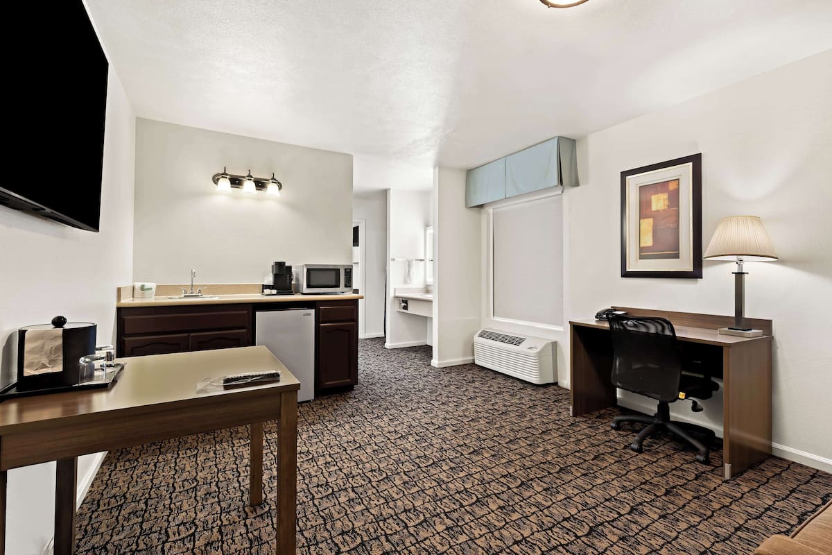 A compact hotel room with a kitchenette, a desk, a chair, a TV, a lamp, and a framed picture on the wall.