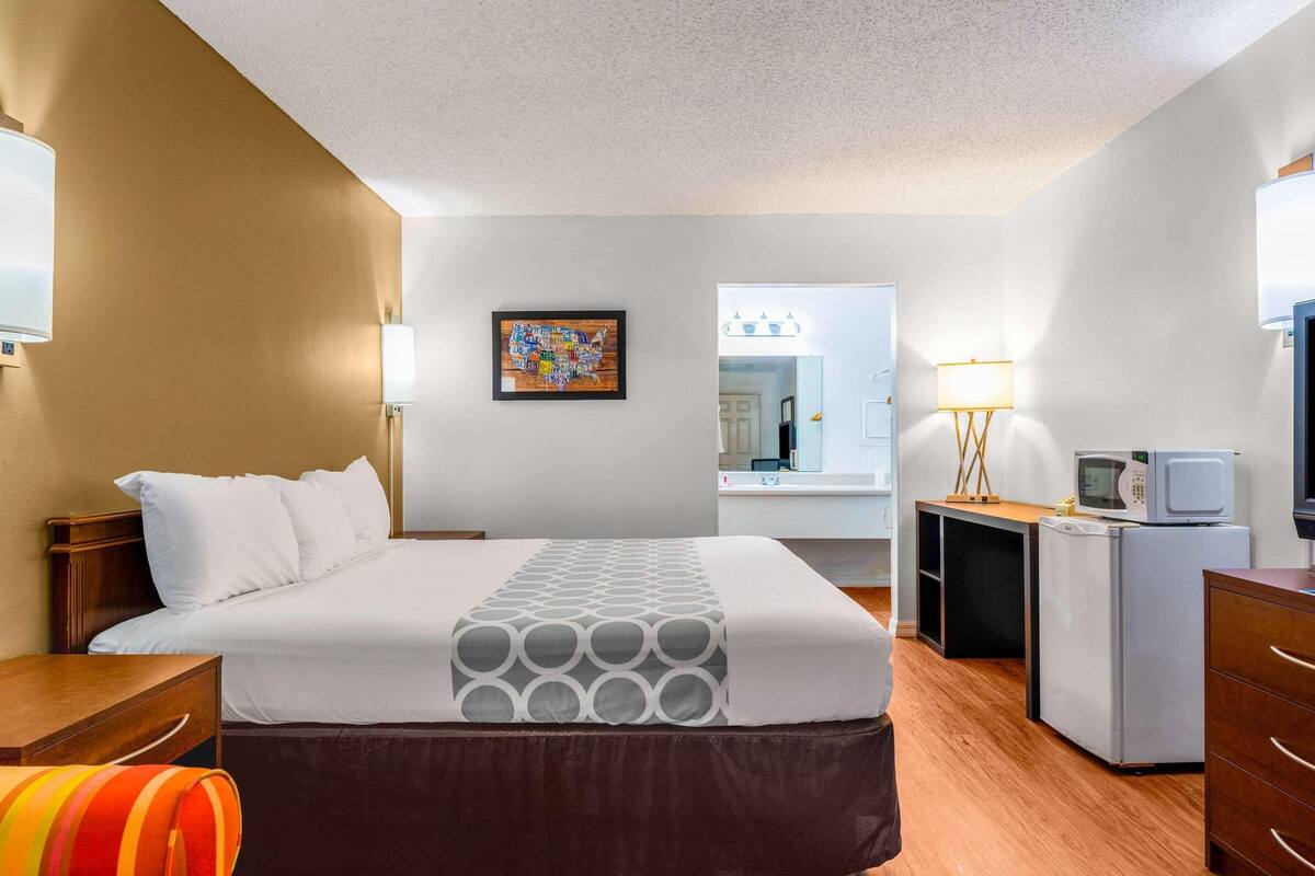 A hotel room with a bed, a desk, a refrigerator, a microwave, a mirror, and a wall art piece.