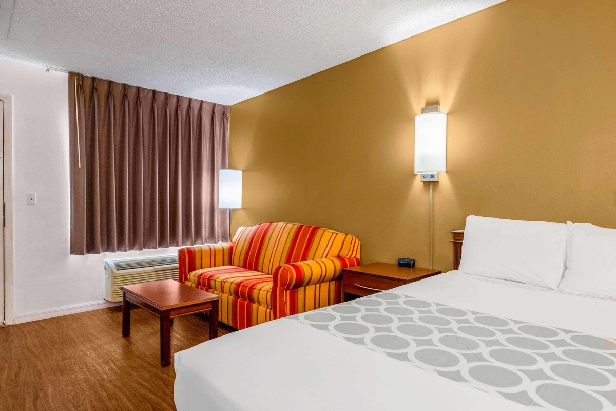 A hotel room with a bed, a striped armchair, a table, a wall-mounted lamp, and a window with curtains.