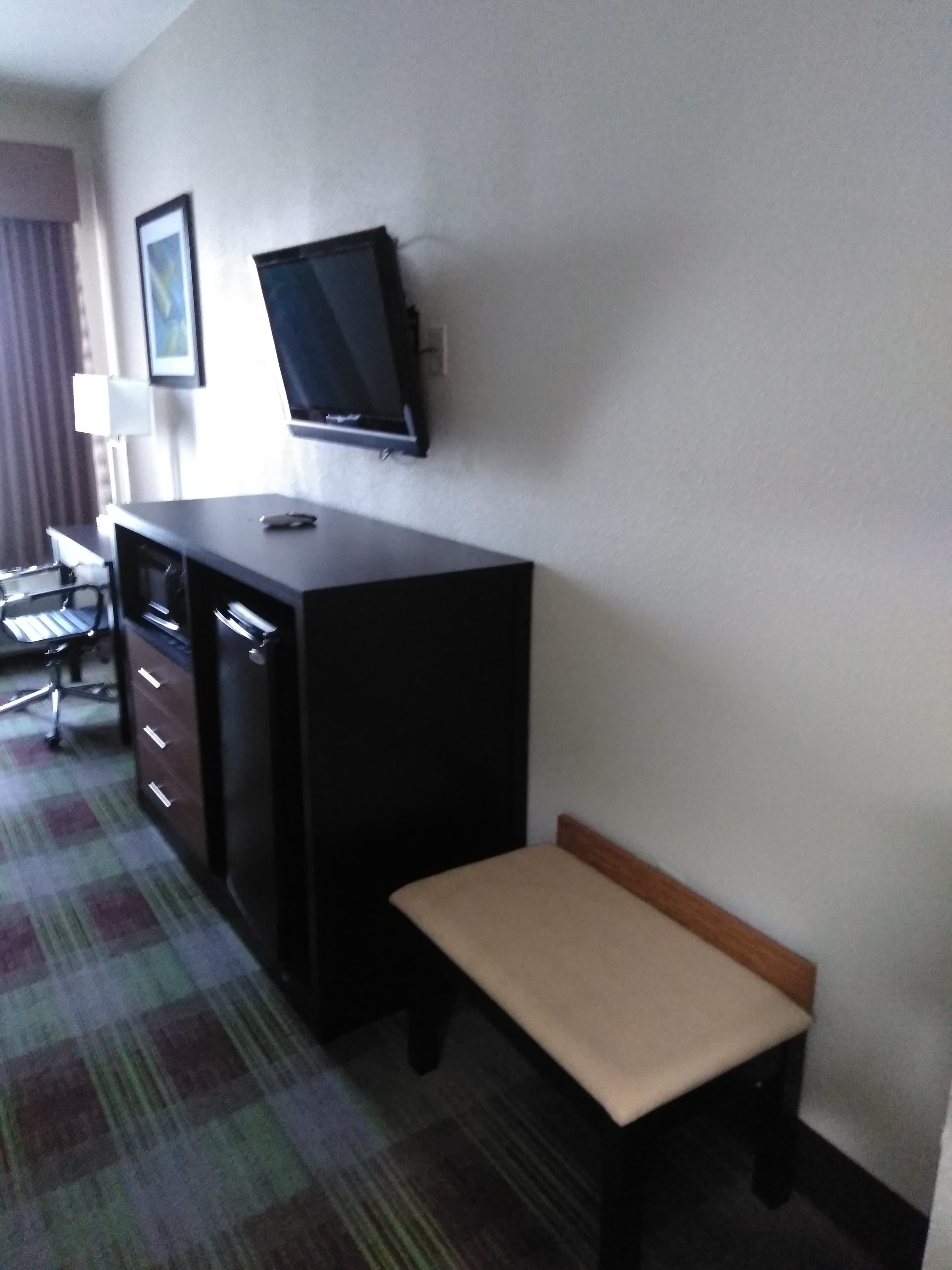 A hotel room with a desk, a television mounted on the wall, a chair, and a bench.