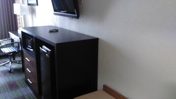 A hotel room with a desk, a television mounted on the wall, a chair, and a bench.