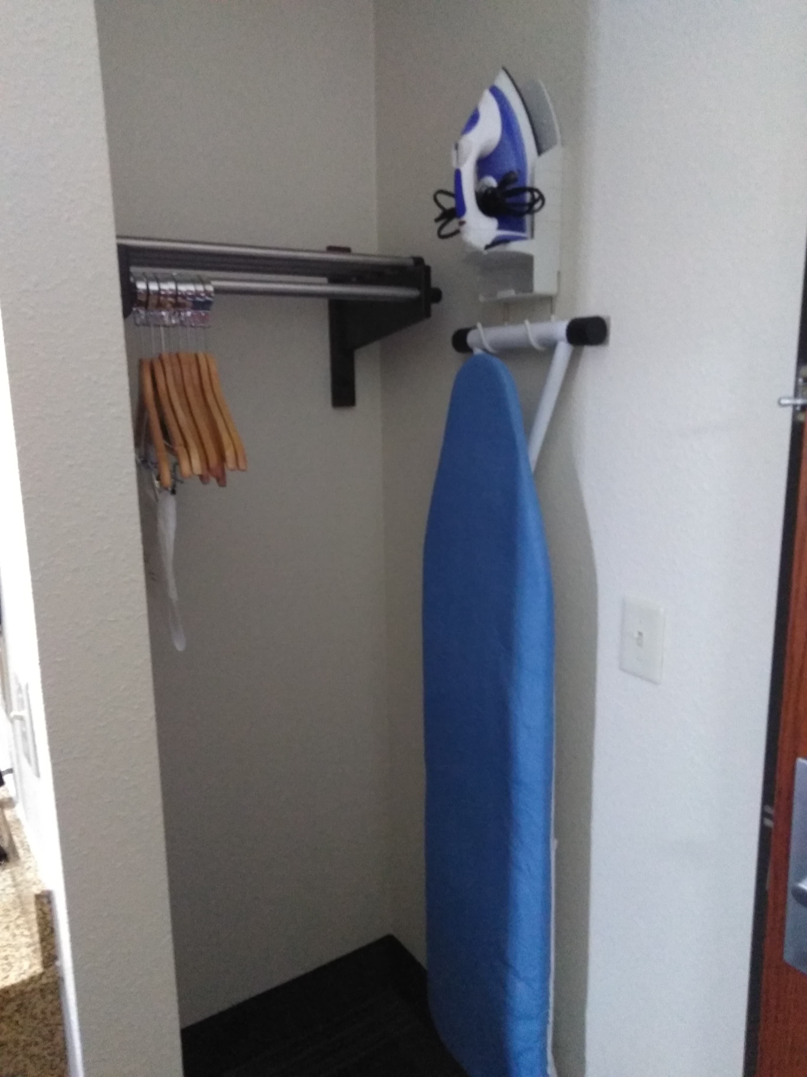 A small room with a wall-mounted ironing board, a hanging rack with clothes, and a door.