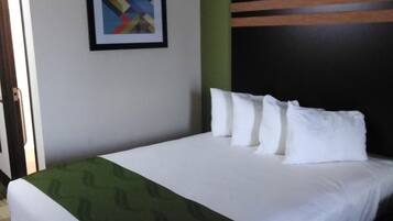 Habitación de hotel con una cama, una colcha estampada verde, almohadas blancas, un cuadro abstracto enmarcado en la pared y una puerta a la izquierda.