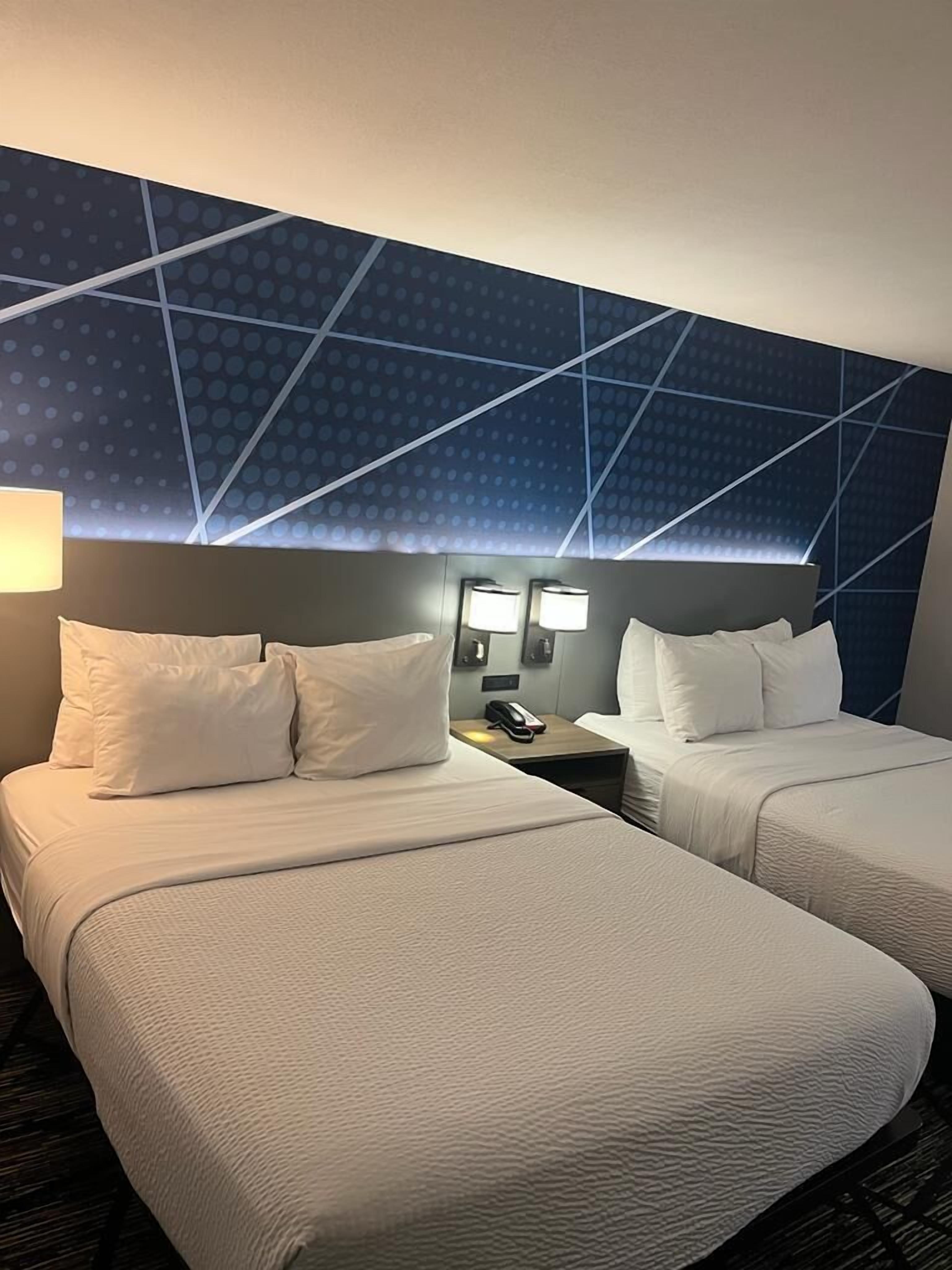 A hotel room with two beds, a nightstand with a phone, and a geometric-patterned ceiling.