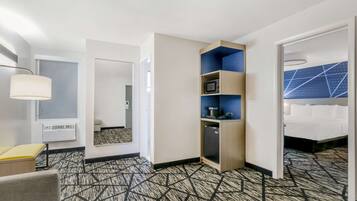 A hotel room with a patterned carpet, a desk with a lamp, a minibar, a bed with white bedding, and a blue accent wall.