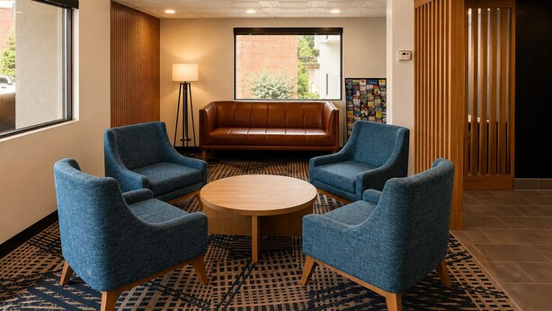 A lobby with a brown leather sofa, four blue armchairs, a round wooden table, patterned carpet, and a large window with a view of a brick building and greenery. - Best Western Plus Atlanta Sandy Springs Perimeter Hotel (Atlanta)