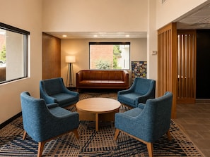 A lobby with a brown leather sofa, four blue armchairs, a round wooden table, patterned carpet, and a large window with a view of a brick building and greenery. - Best Western Plus Atlanta Sandy Springs Perimeter Hotel (Atlanta)