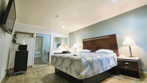 A hotel room with a bed, bedside lamps, a nightstand, a desk with a lamp, a mirror, and a wardrobe. - Welcome Inn (Corona)