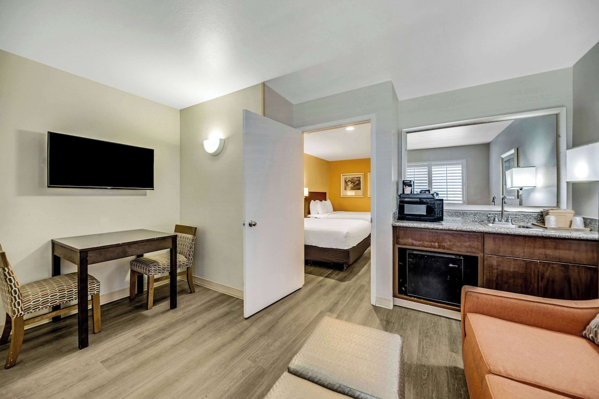 Quarto de hotel com televisão de tela plana, mesa de jantar com cadeiras, sofá, área de cozinha com micro-ondas e uma cama no quarto ao lado.