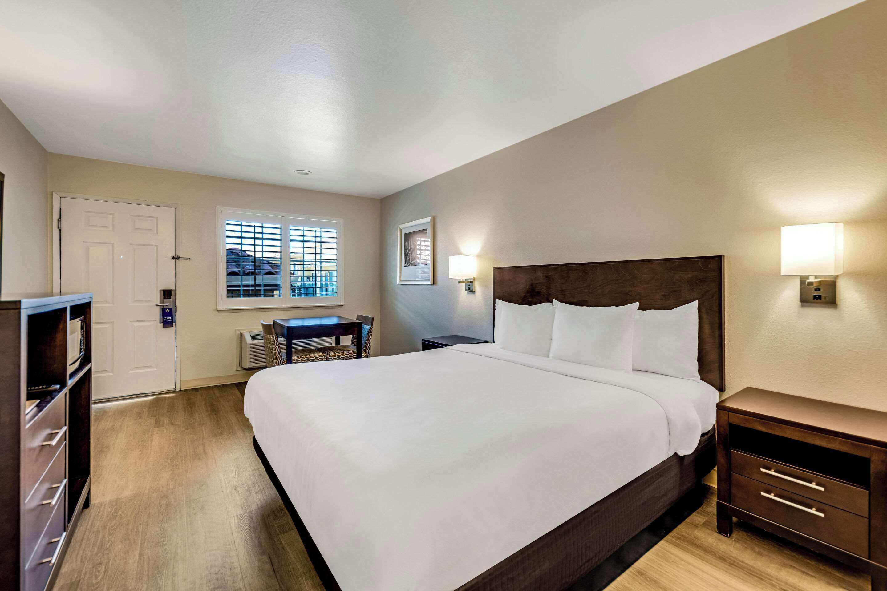 A hotel room with a large bed, a desk, a chair, a wardrobe, and a window with blinds.