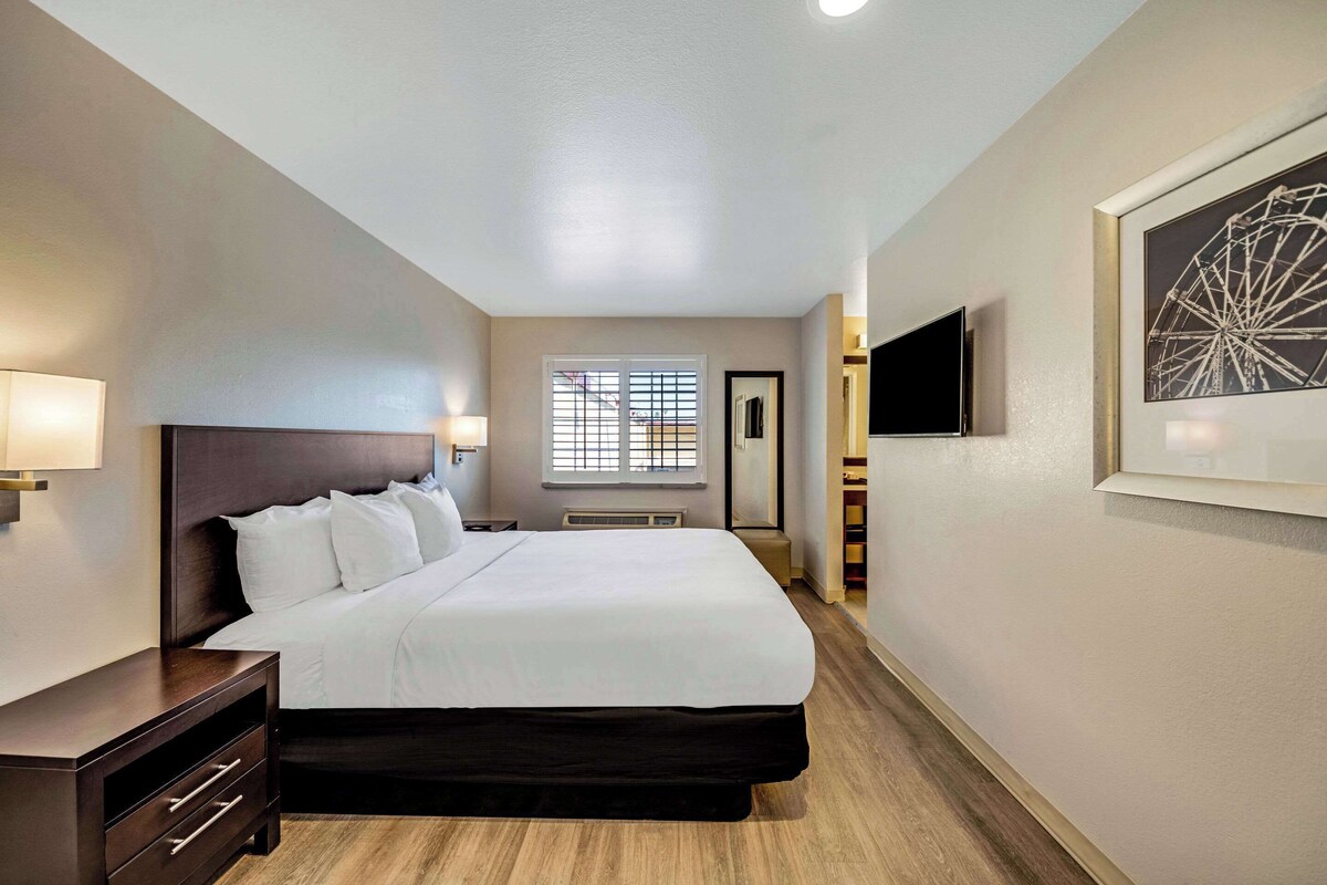 Quarto de hotel com cama grande, luminárias laterais, janela com persianas, televisão montada e quadro na parede.