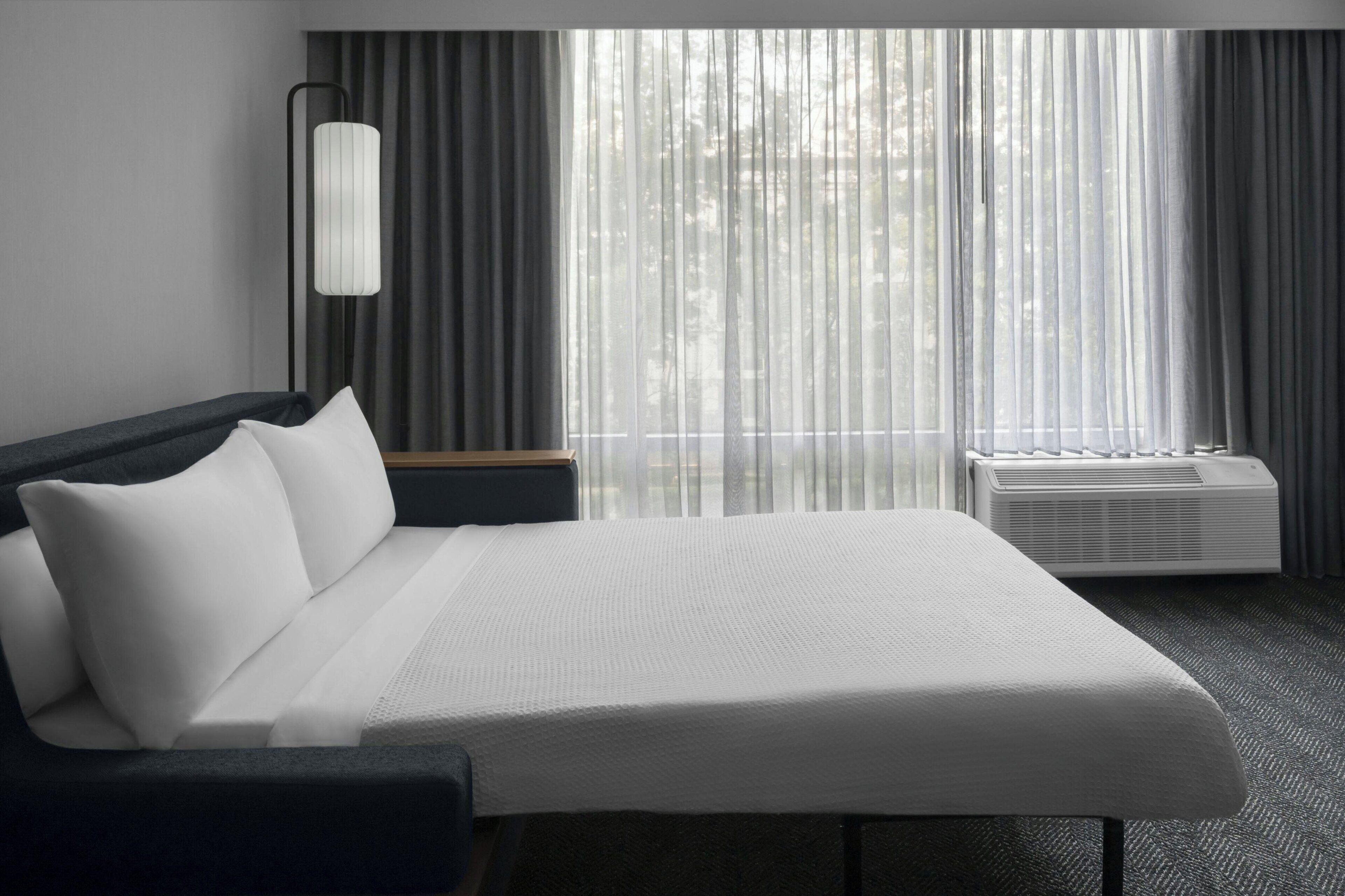 A modern hotel room with a large bed, a bedside table, a lamp, and a window with curtains.