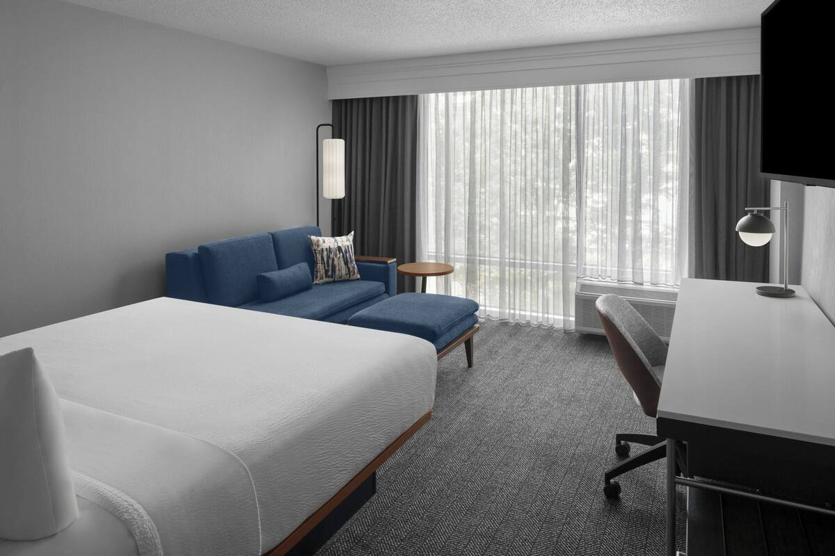 A hotel room with a large bed, a sofa, a desk, and a chair. There is a window with curtains, a flat-screen TV, and a lamp.