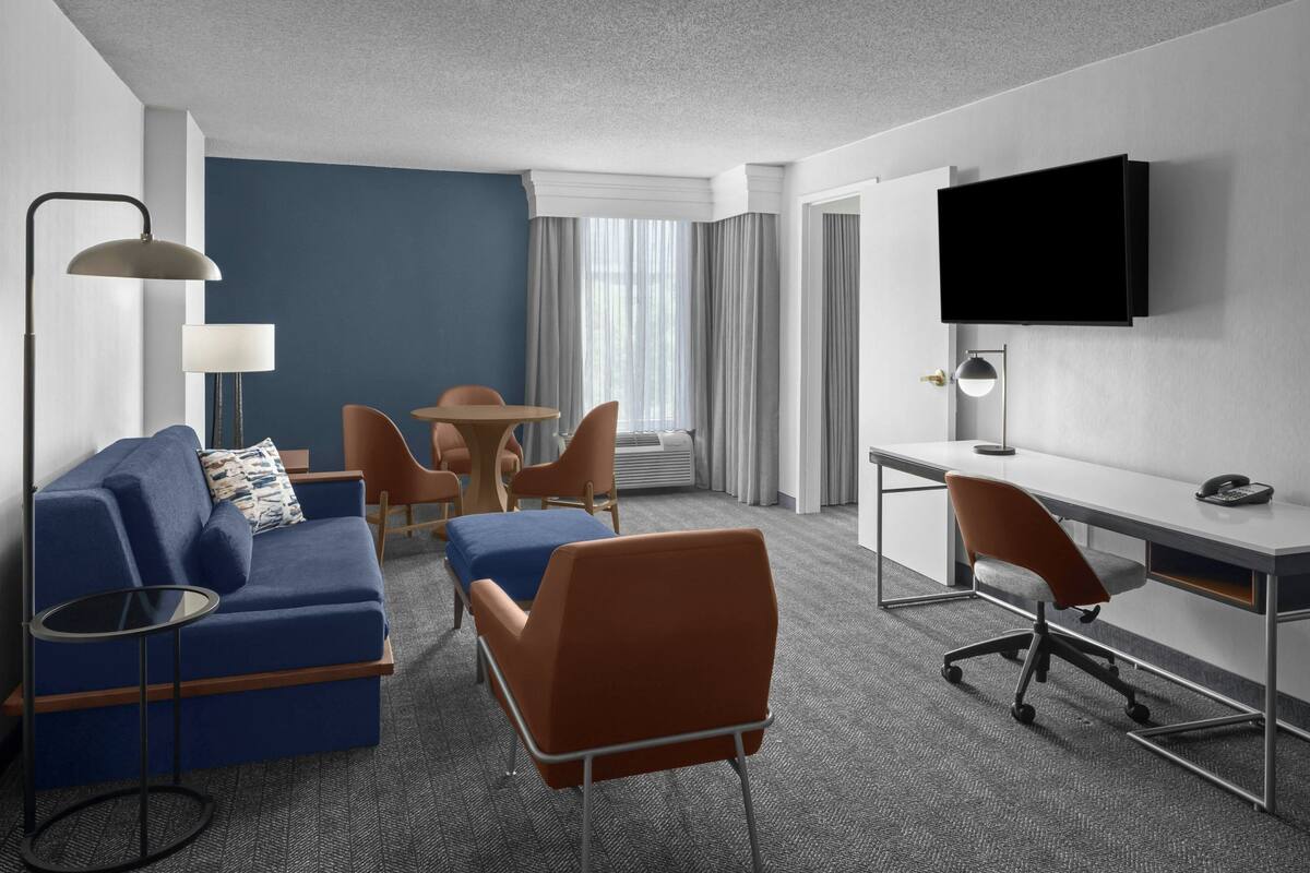 A hotel room with a blue sofa, orange chairs, a desk, and a TV mounted on the wall.