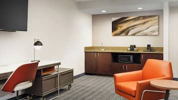 A hotel room with a kitchenette, a flat-screen TV, a desk with a chair, and an orange armchair.