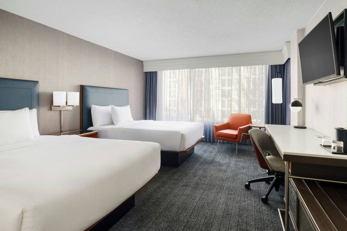 A hotel room with two beds, a desk, a chair, a TV, and a large window with curtains.