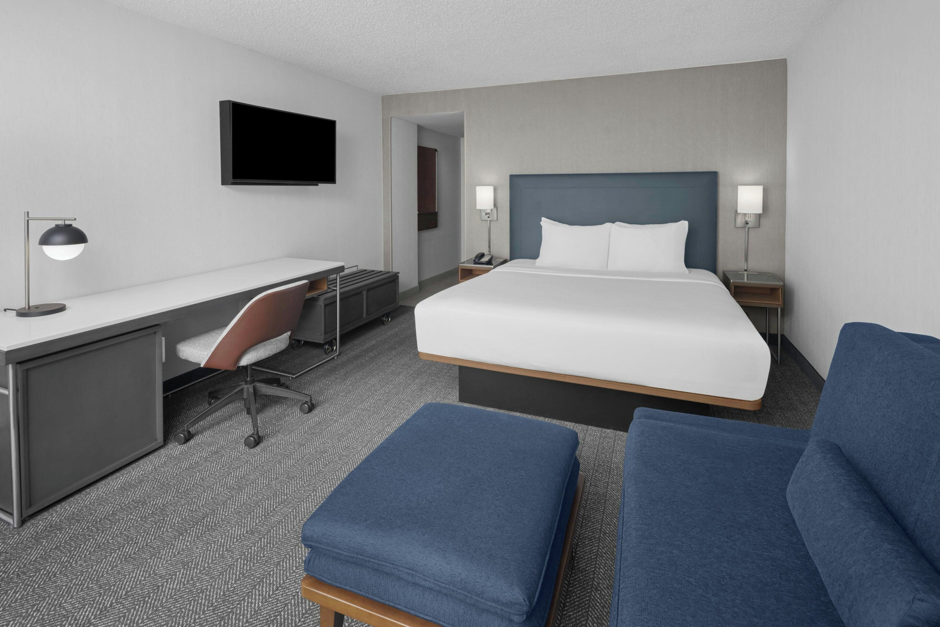 Una habitación de hotel moderna con una cama grande, un escritorio con silla, un televisor montado en la pared y una mesita de noche con lámpara.