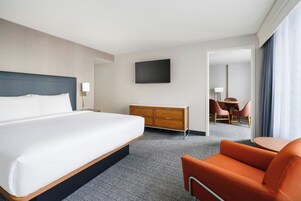 A hotel room with a large bed, a wooden dresser, a flat-screen TV, and a seating area with a chair and a small table.