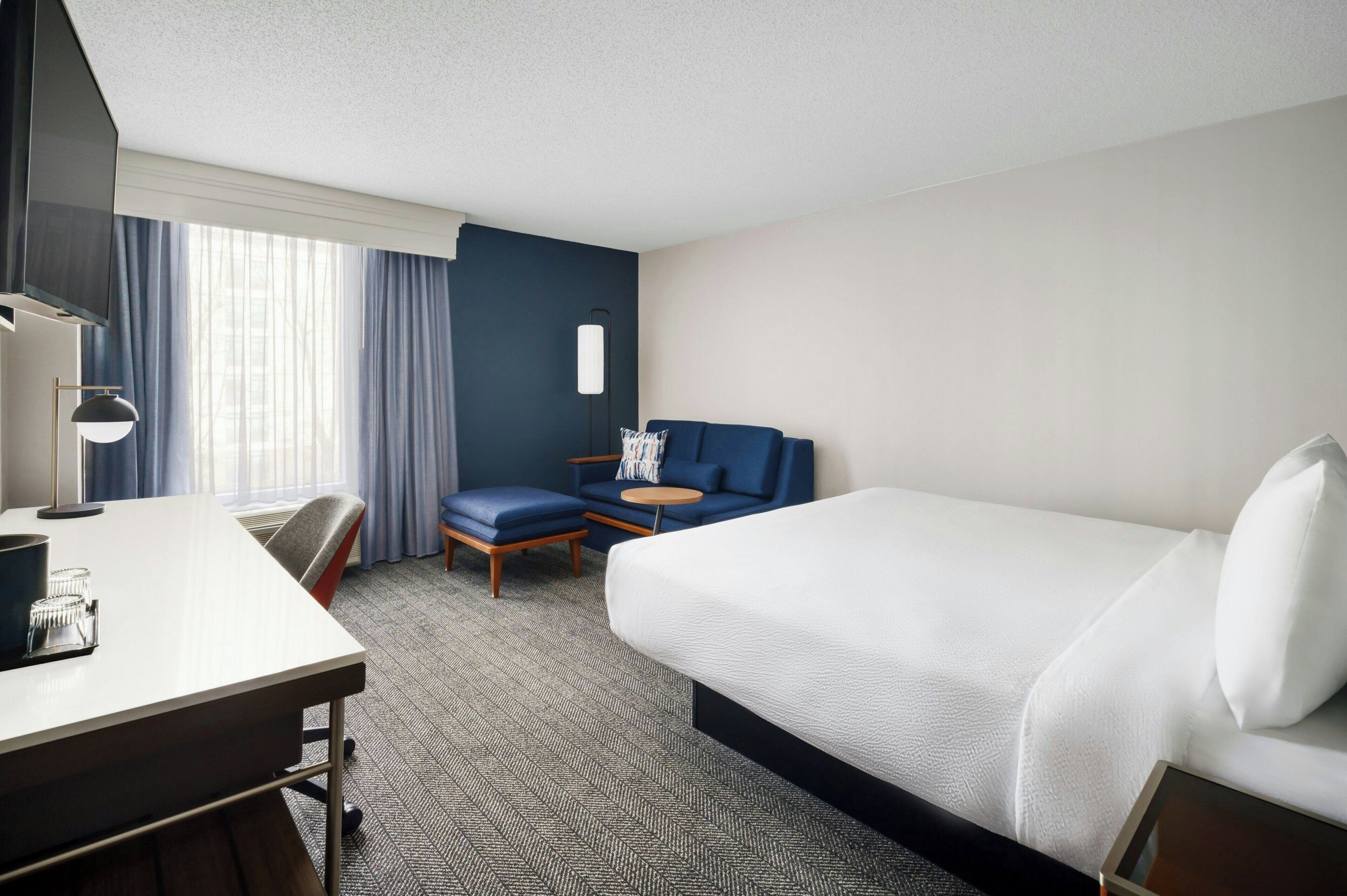 A hotel room with a large bed, a desk with a lamp, a chair, and a small table.
