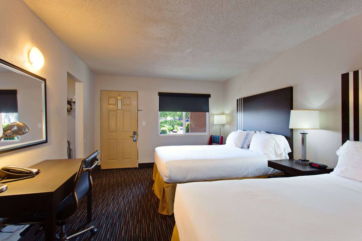 Une chambre d’hôtel avec deux lits, un bureau avec un téléphone, une chaise, une porte et une fenêtre avec des stores.
