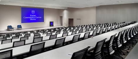 A conference room with rows of tables and chairs, a stage with a screen, and a door in the background.