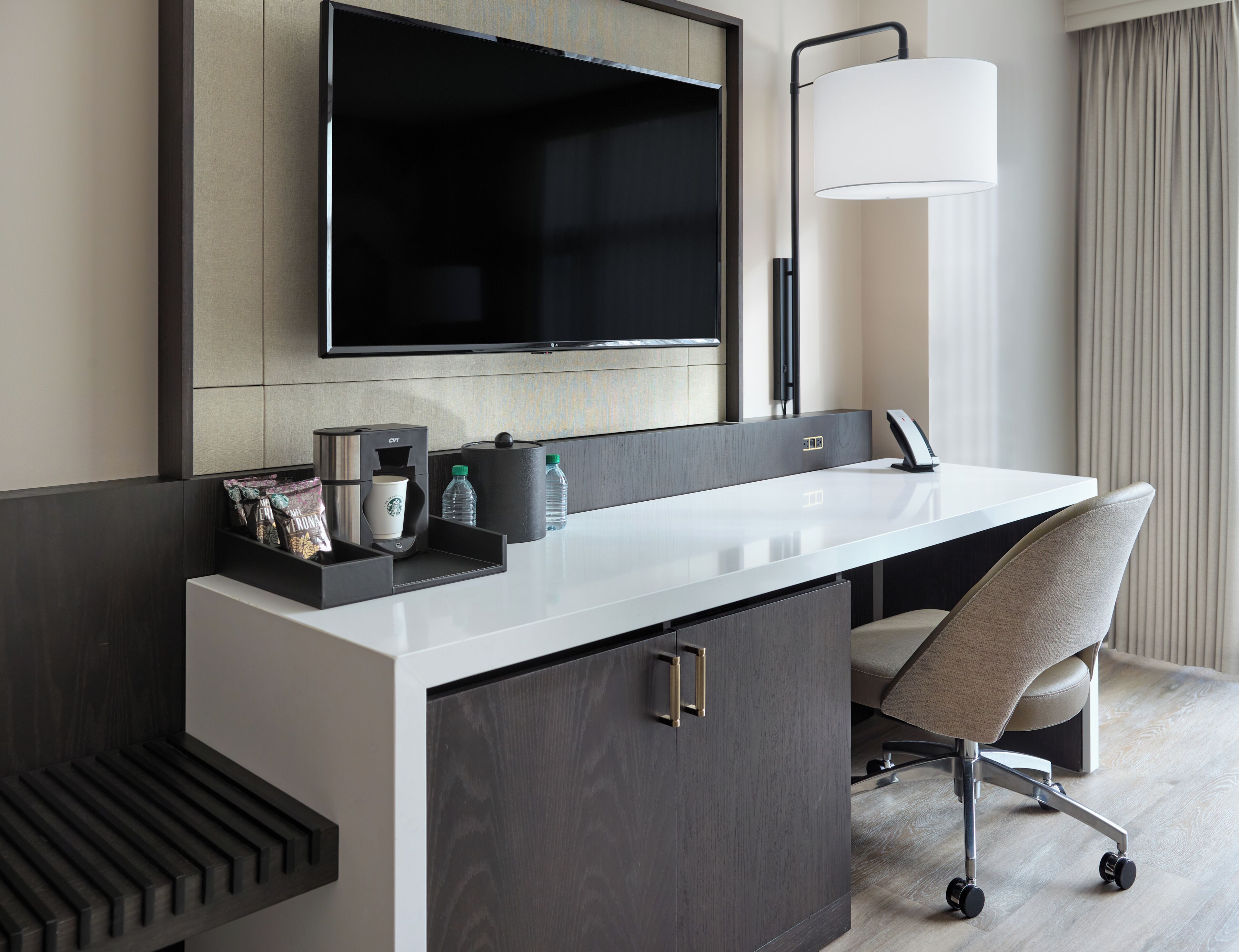 Habitación de hotel moderna con una gran televisión de pantalla plana sobre un tocador blanco, un escritorio con una silla y una lámpara de pie grande.