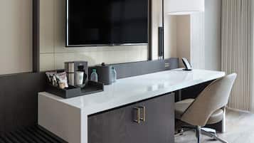 Habitación de hotel moderna con una gran televisión de pantalla plana sobre un tocador blanco, un escritorio con una silla y una lámpara de pie grande.