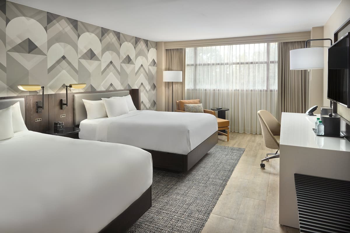 A hotel room with two beds, a desk, a chair, a TV, and a large window with curtains.