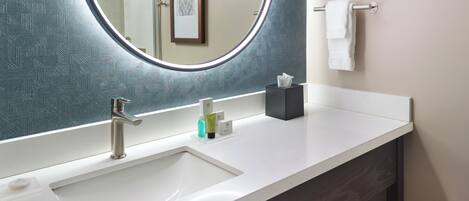 A bathroom with a white sink, a mirror with a light, a towel rack, and a framed picture on the wall.
