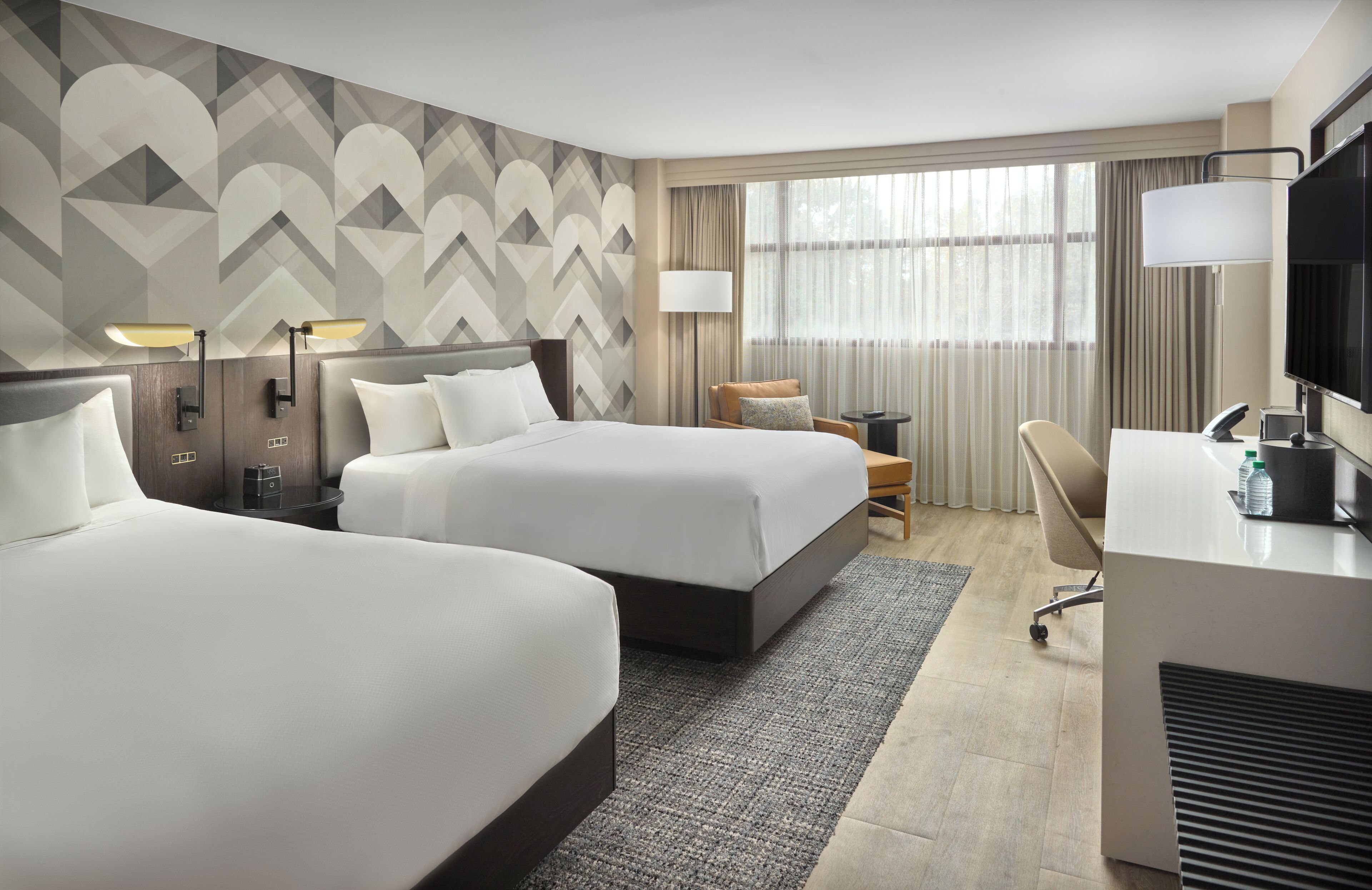 A hotel room with two beds, a desk, a chair, a TV, and a window with curtains.