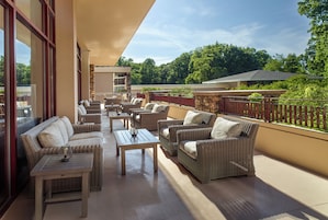 A covered outdoor seating area with wicker furniture and tables, a stone wall, and a view of the surrounding greenery.