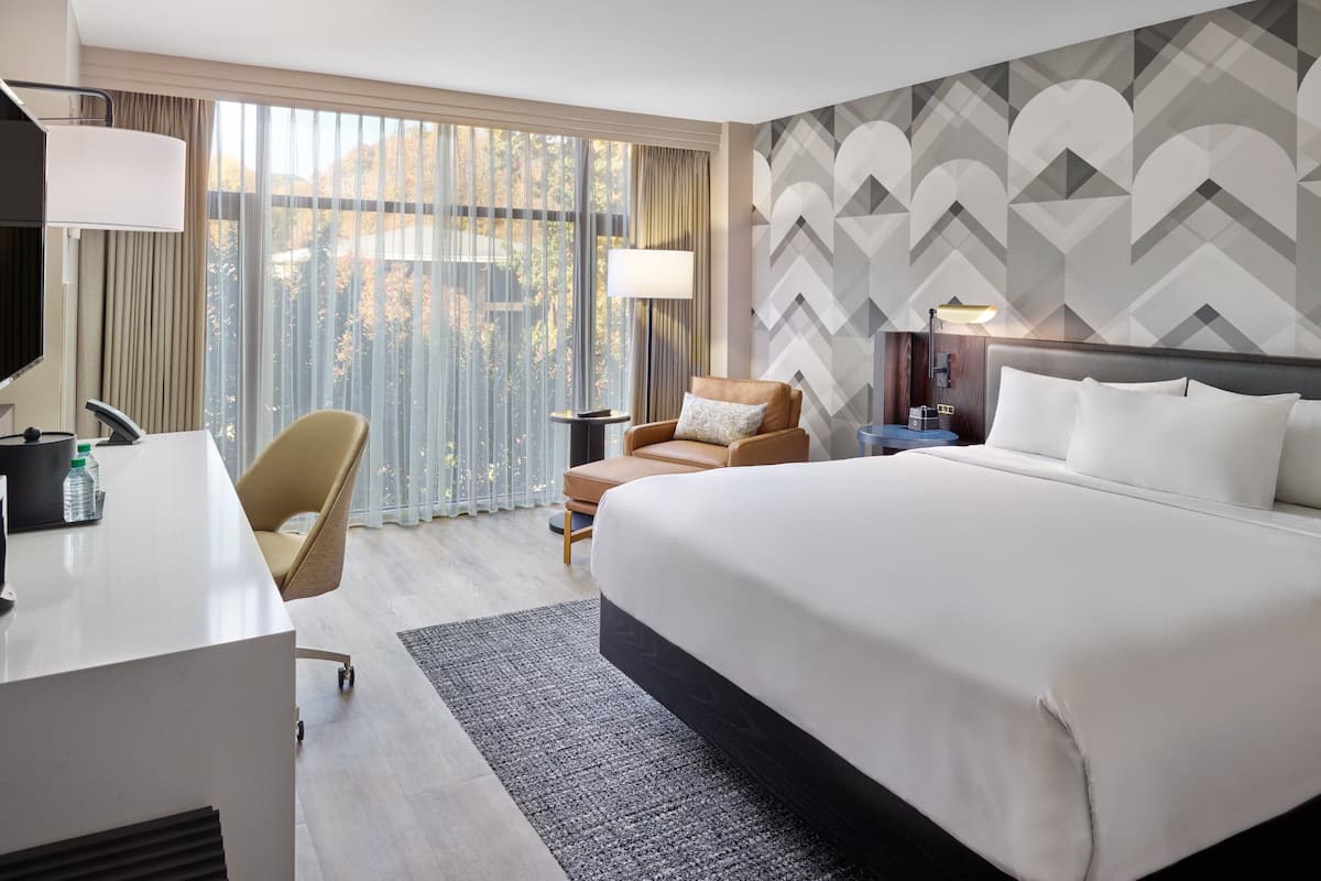 A hotel room with a large bed, a desk, a chair, and a view of the outdoors through large windows.