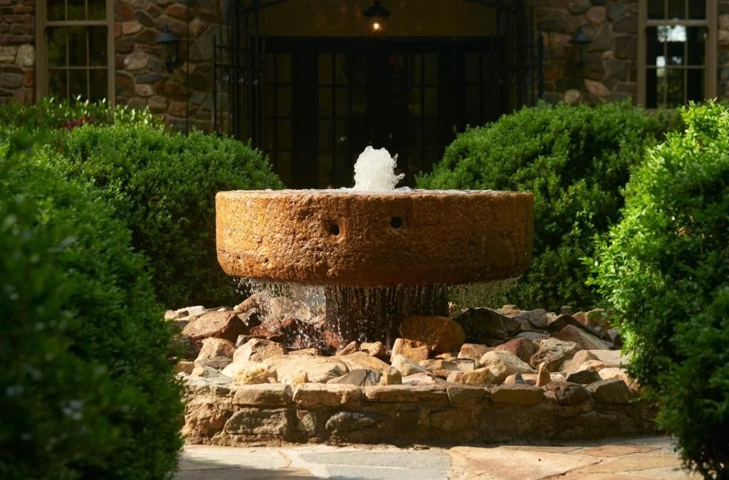 A stone fountain with water spouting from the center, surrounded by neatly trimmed bushes and a stone base.
