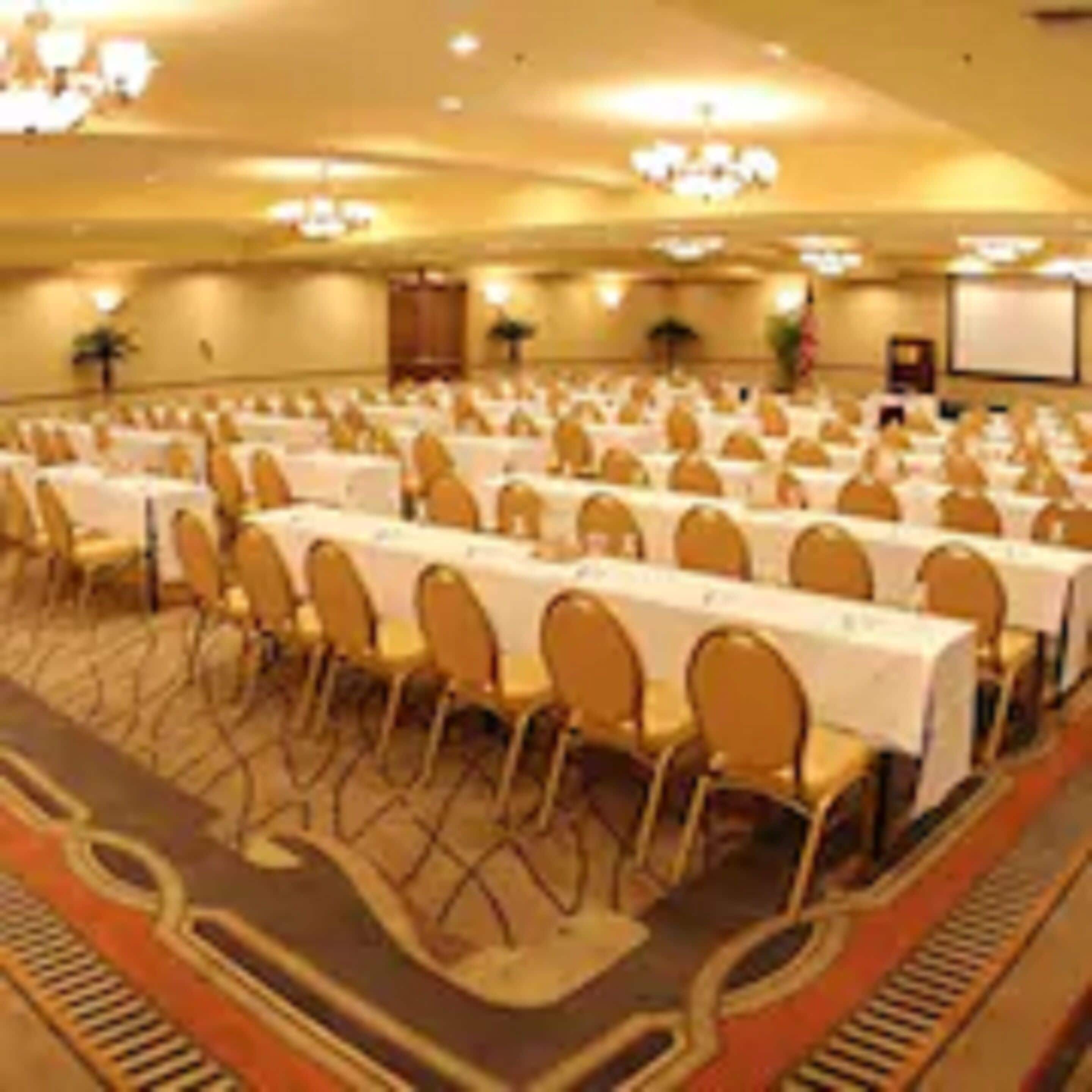 Un salón de conferencias con filas de mesas y sillas, una pantalla de proyector y un atril.