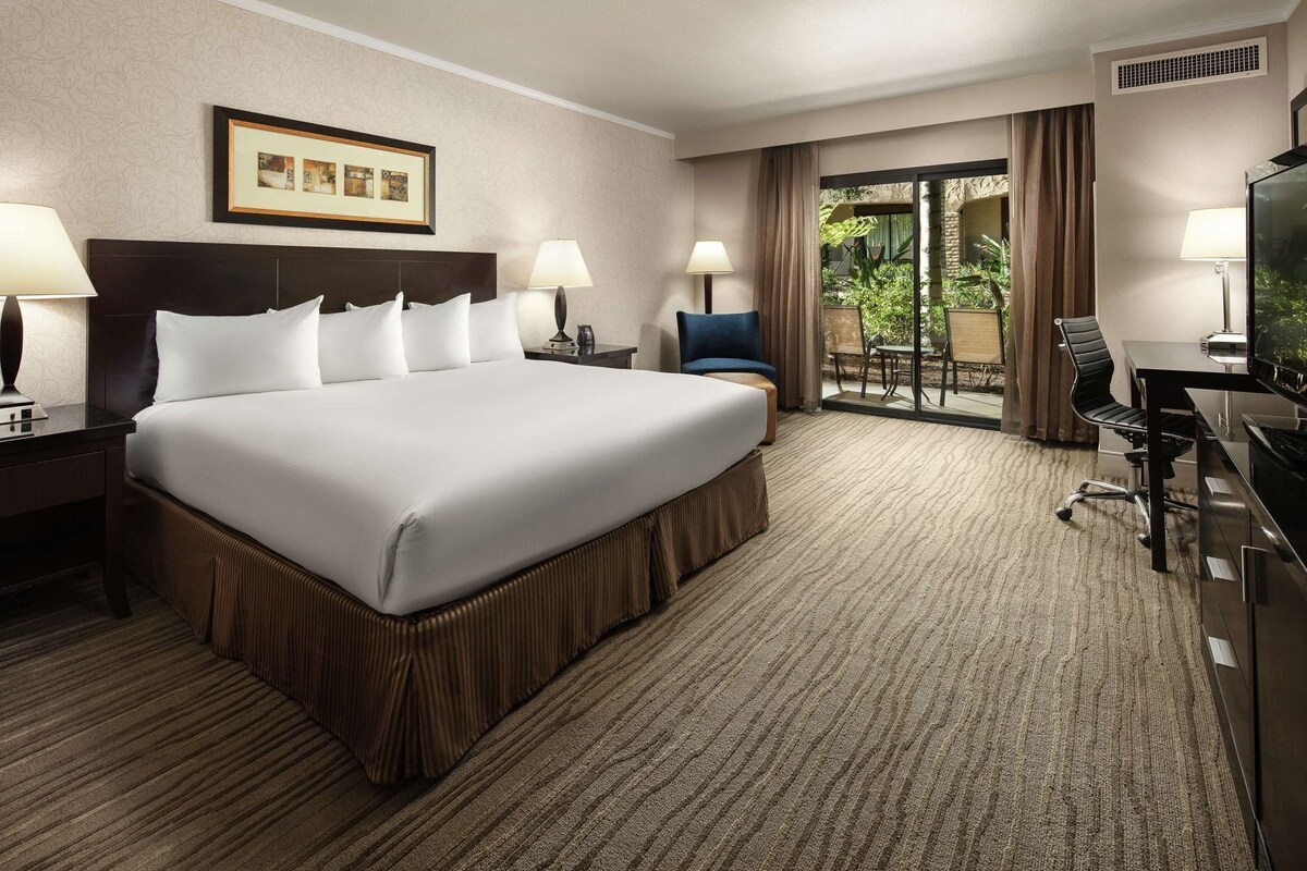 A hotel room with a large bed, two bedside tables with lamps, a desk with a chair, a television, and a view of greenery through the window.