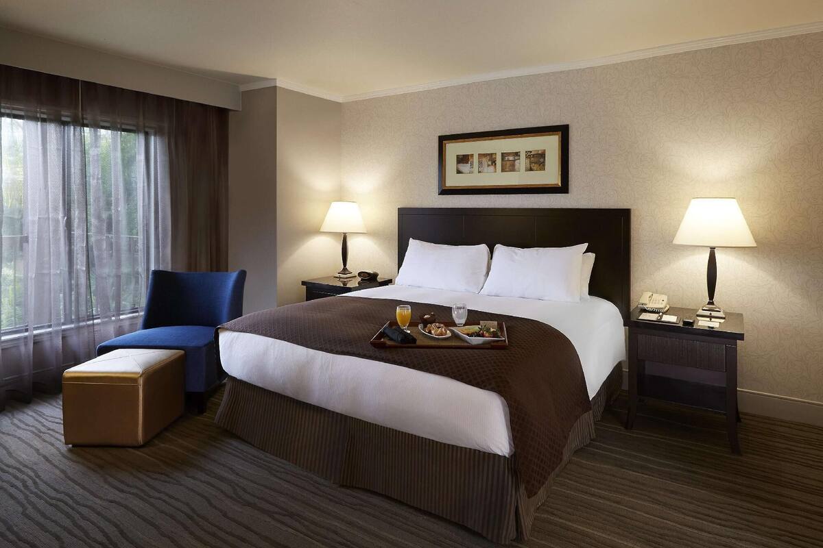 A hotel room with a large bed, two bedside tables, a chair, a small ottoman, and a window with curtains.