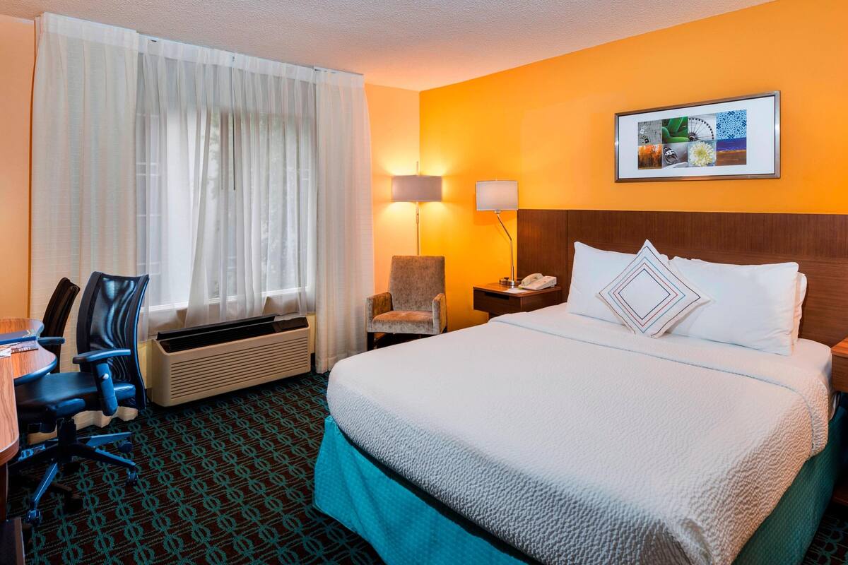 A hotel room with a bed, a desk, a chair, a lamp, and a picture on the wall.