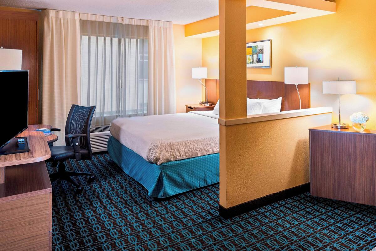 A hotel room with a bed, desk, chair, and a large window with curtains.