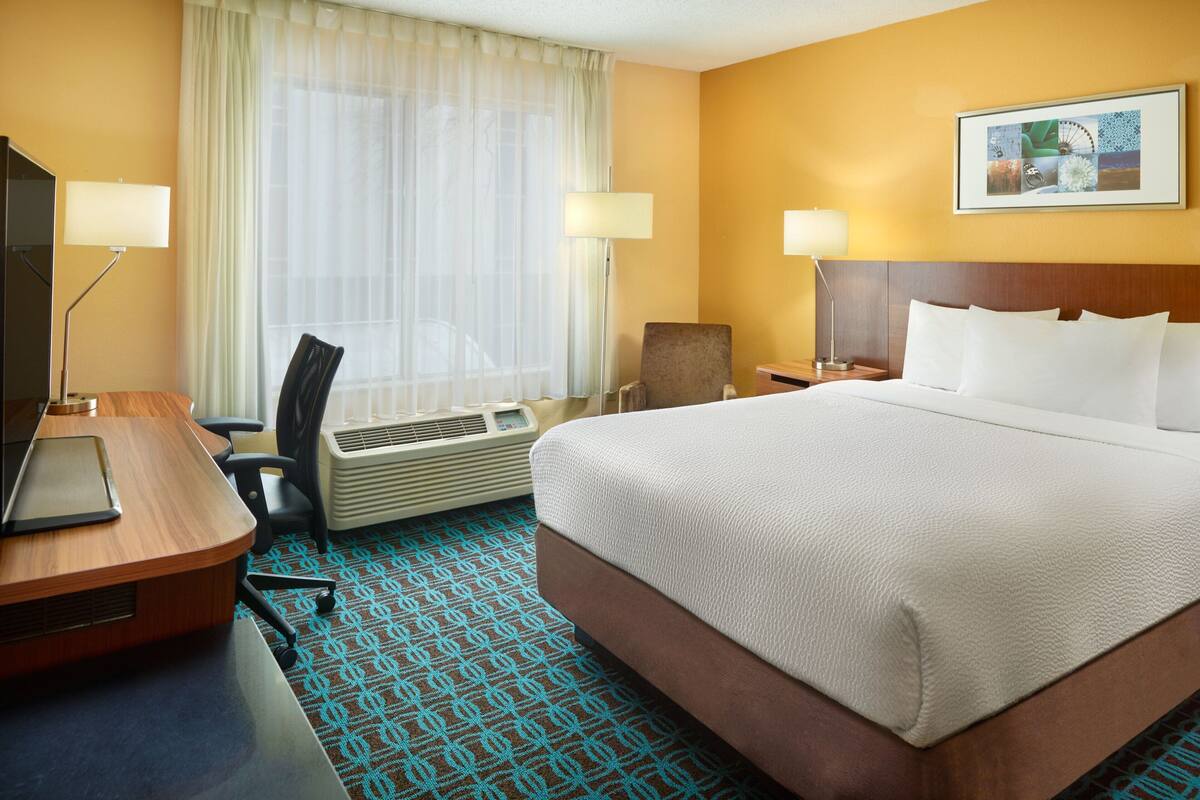 A hotel room with a large bed, a desk with a computer, a chair, a desk lamp, and a wall-mounted picture.